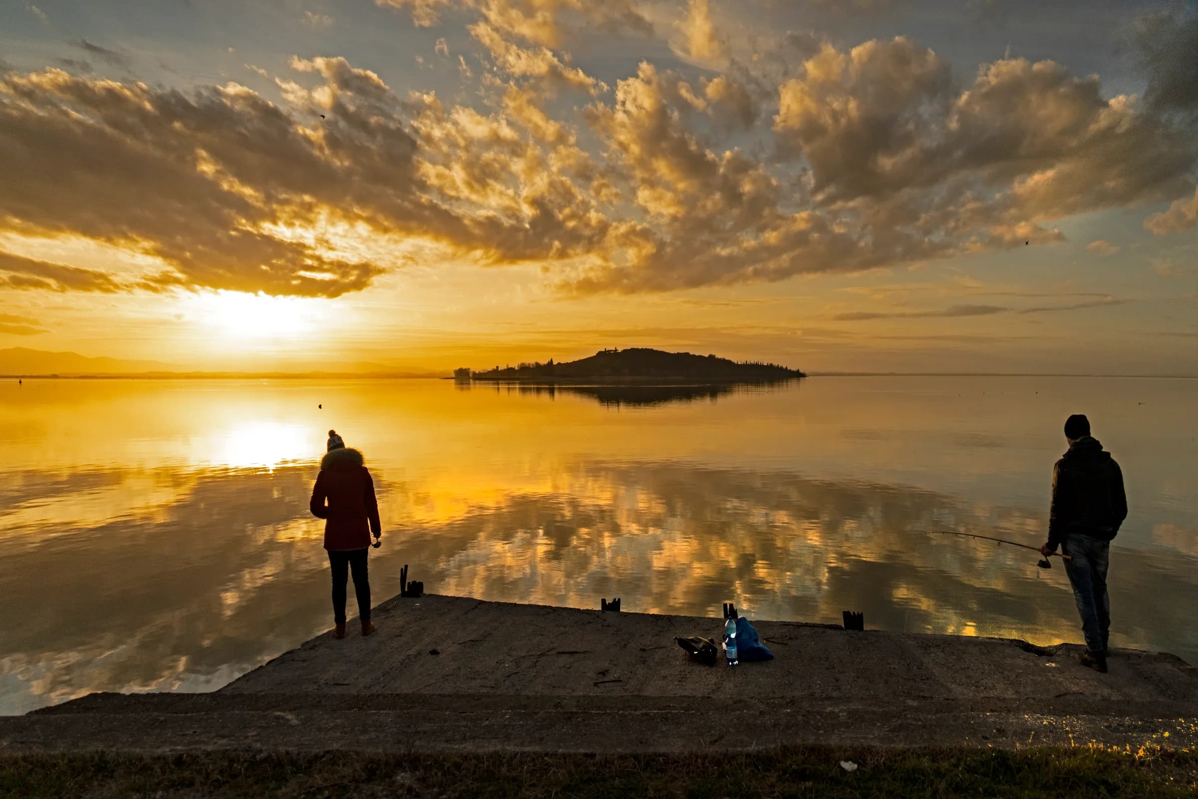 Lake Trasimeno at sunset in winter, with Isola Polvese on the horizon and two people fishing from the jetty