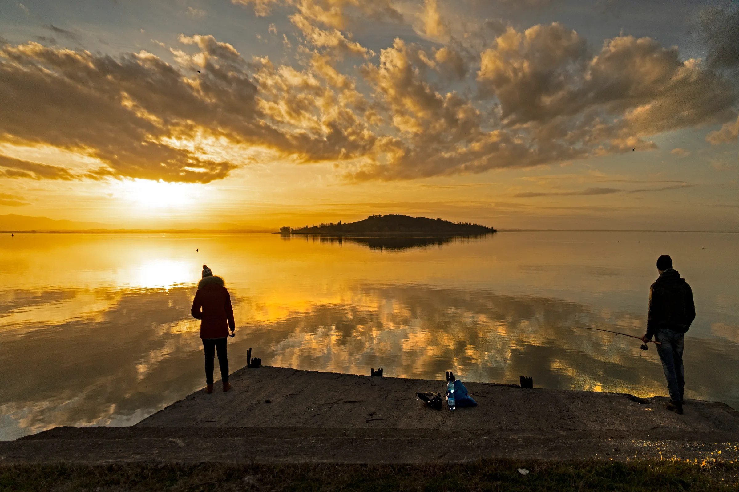 Il lago Trasimeno al tramonto d'inverno, con l'Isola Polvese all'orizzonte e due pescatori sul molo